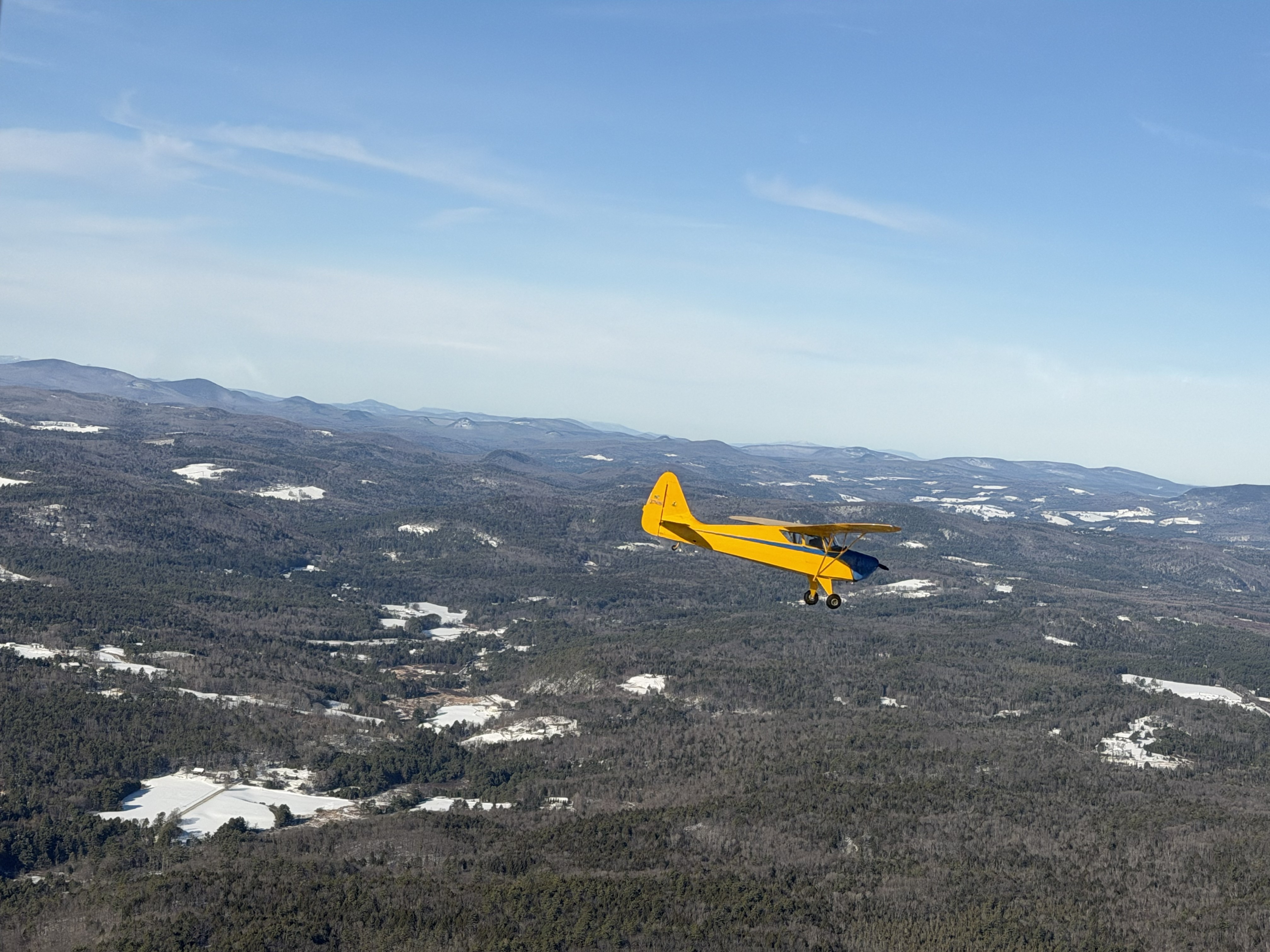 Yellow Piper Cub flying over snow-covered Vermont mountains on a clear winter day